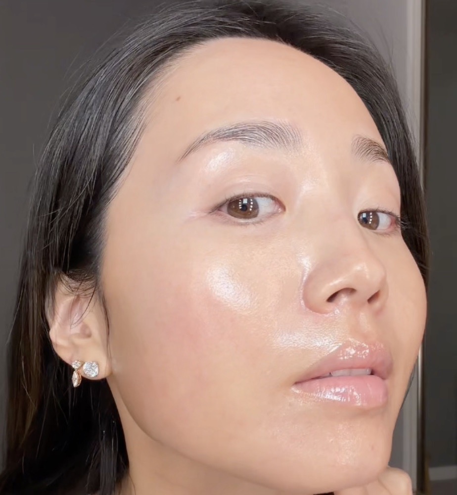 A close-up of a woman with dark hair and dewy, glowing skin looking toward the camera.