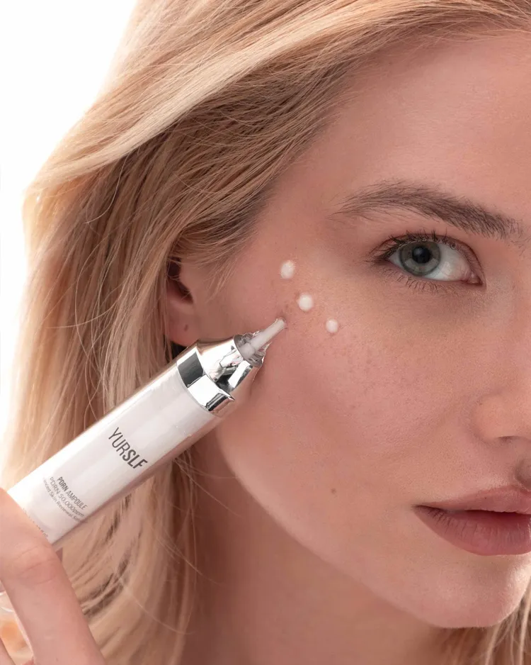 A woman applies white cream under her eye from a syringe-like applicator.