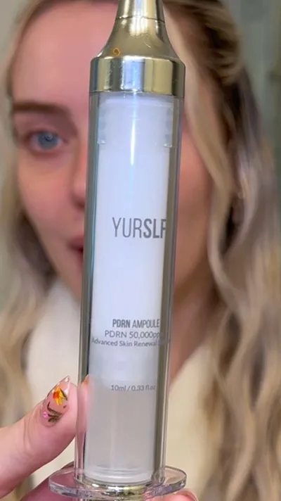 A woman with floral nails holds a syringe-like dispenser of YURSLA PDRN Ampoule skin serum.