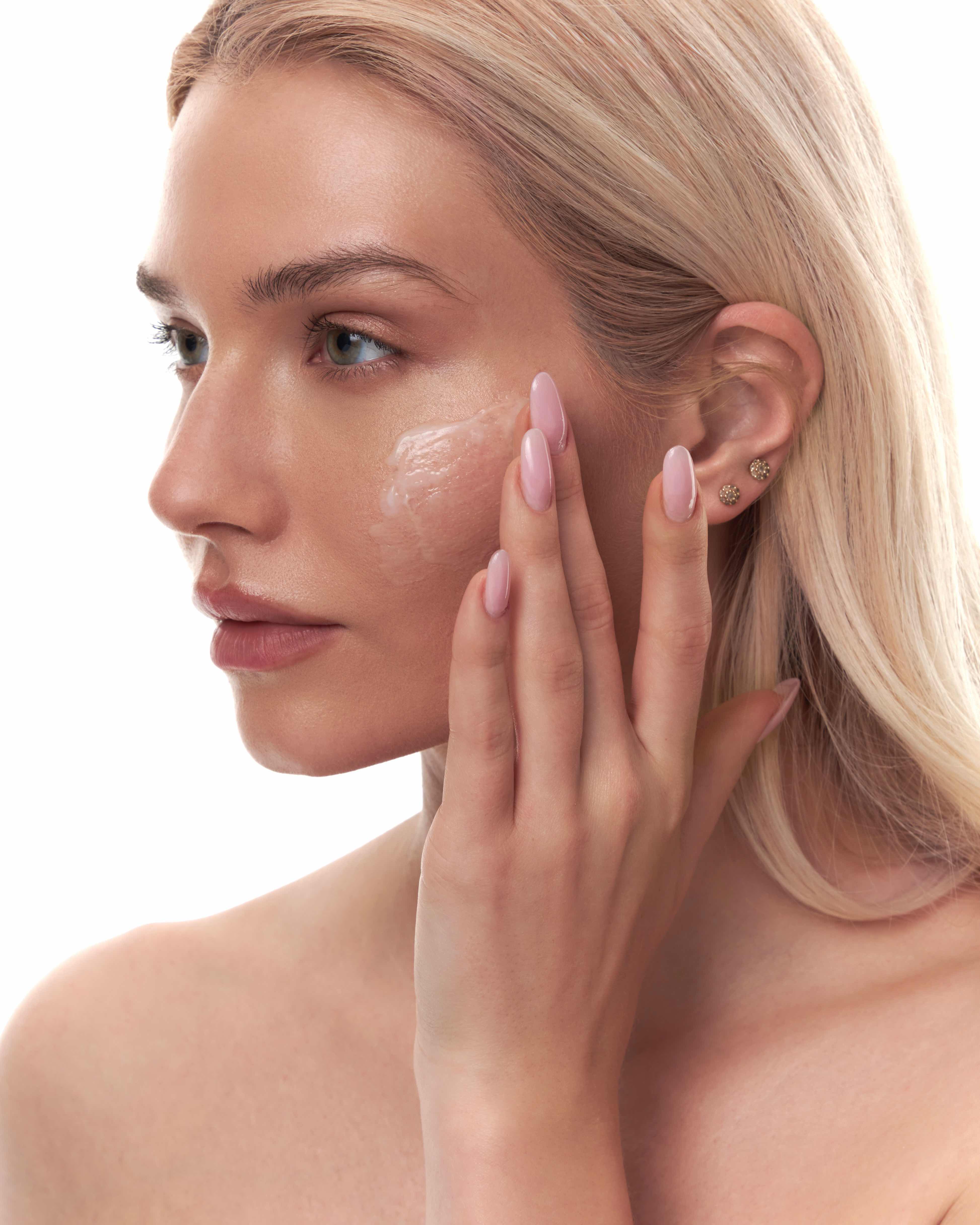A blonde woman in profile applies a cream to her face against a white background.
