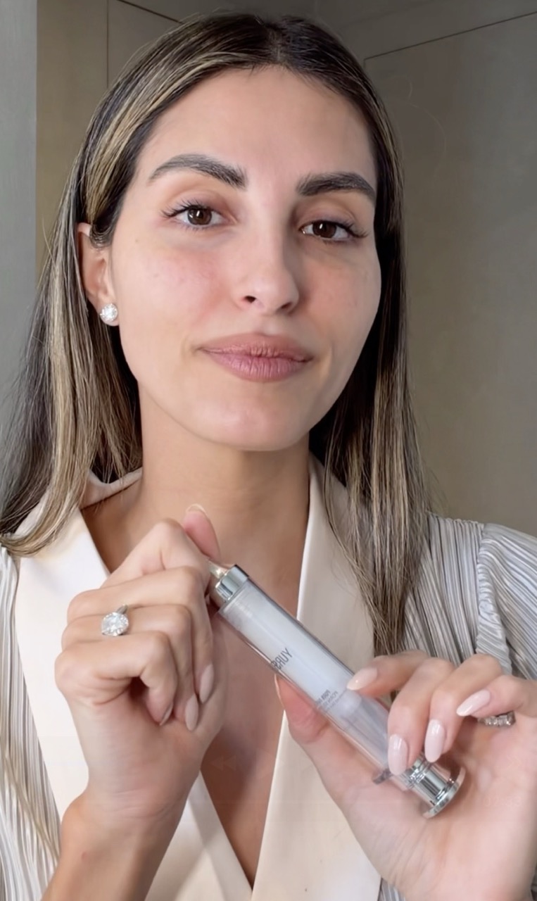 A woman with long highlighted hair holds a skincare product packaged in a syringe-style applicator.