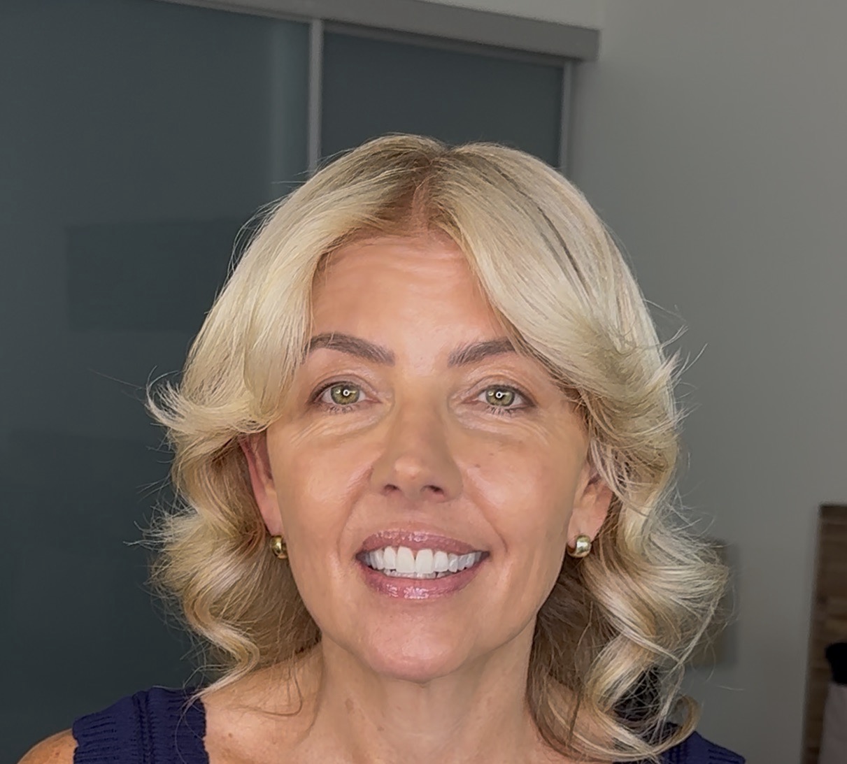 A close-up of a smiling woman with blonde, shoulder-length hair and green eyes, wearing a blue top.
