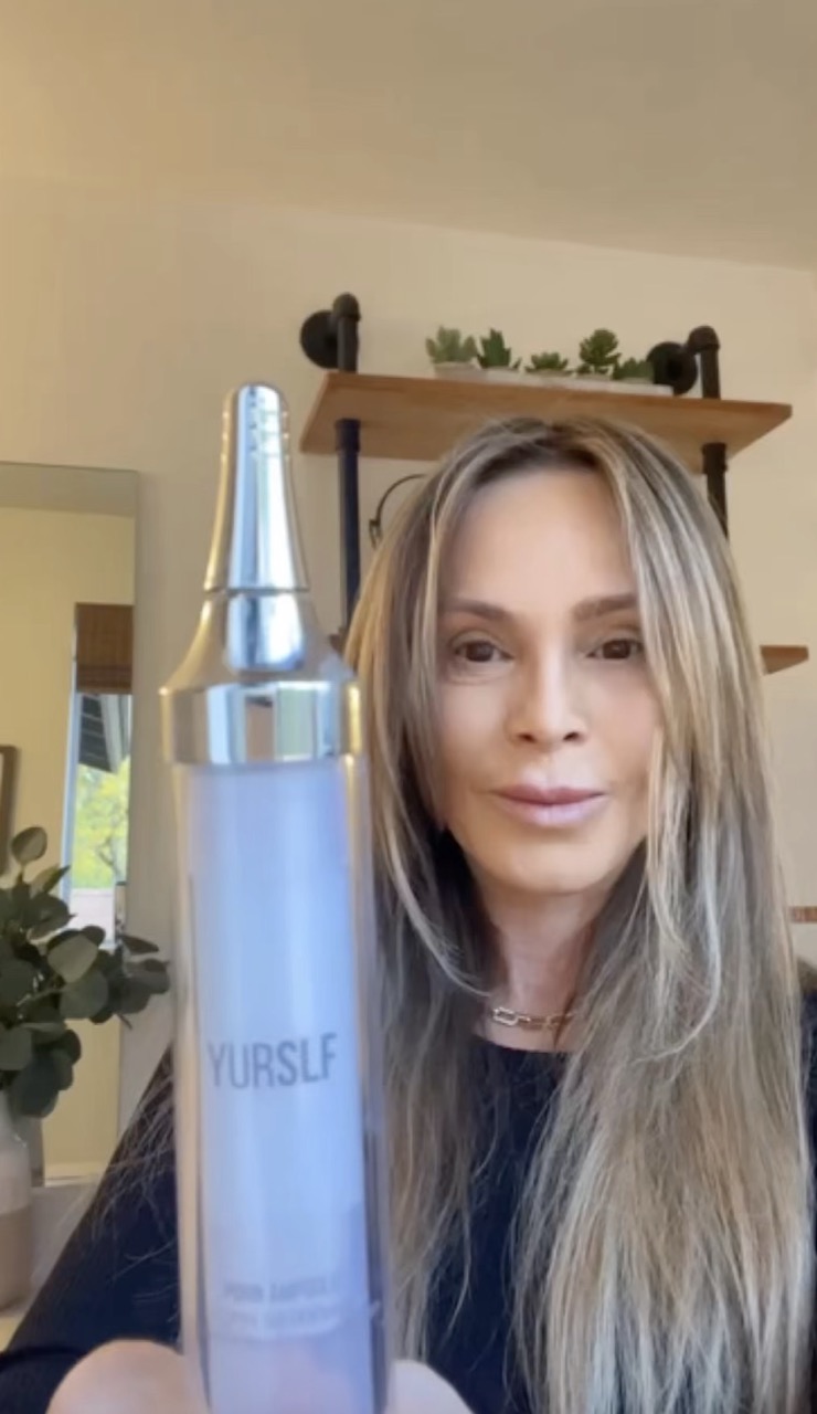 A woman with long highlighted hair holds up a bottle of YURSLF skincare product.