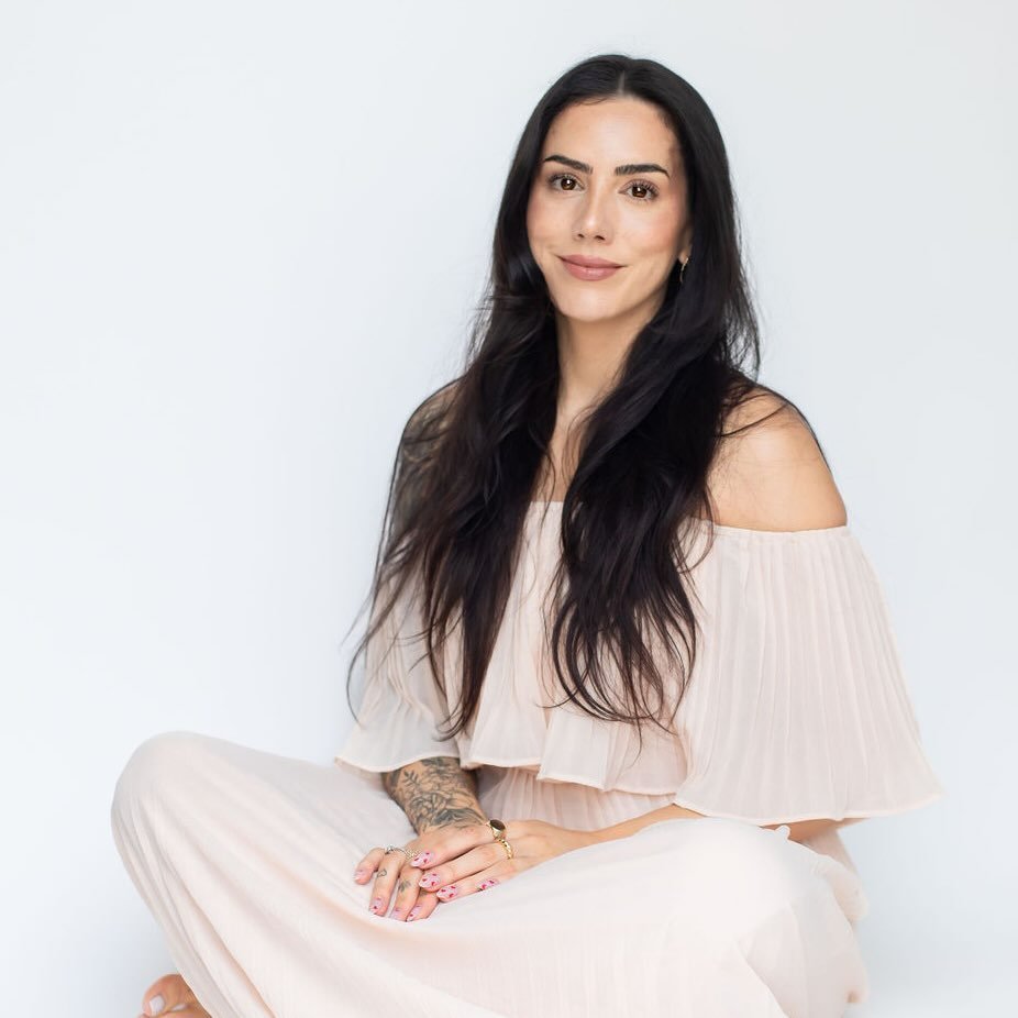A woman with long, dark hair and tattoos sits cross-legged against a white background, smiling at the camera.