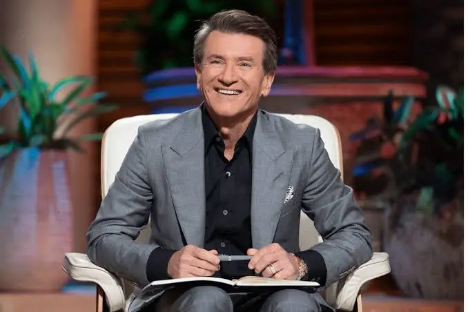 A smiling man in a grey suit and black shirt sits in a chair with a notebook.