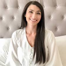 Smiling woman with long dark hair sits on a bed with a tufted headboard.