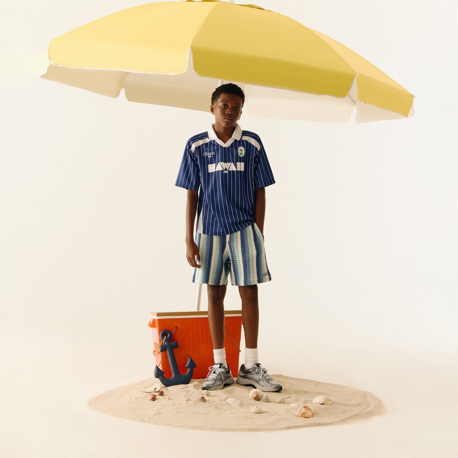 Person standing on sand under a yellow umbrella with a cooler and anchor.