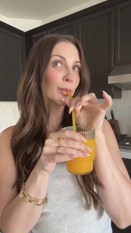 Person drinking orange juice with a straw in a kitchen.