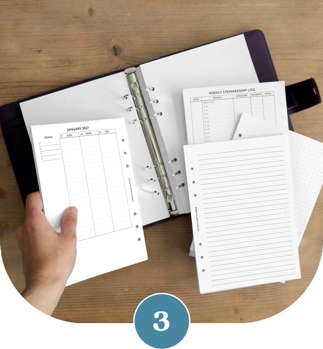 A person holds planner inserts, including a calendar and log, over an open six-ring binder on a wood surface.