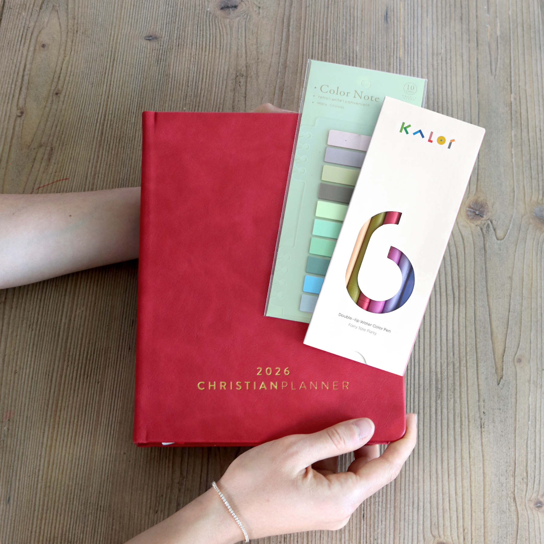 Hands holding a 2026 Christian Planner, color notes, and a pen set on a wooden table.