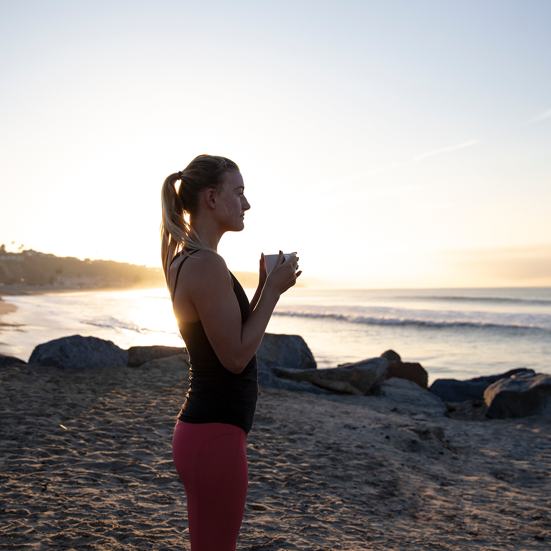 A woman in athletic wear holds a mug and looks out at the ocean from a beach at sunrise.