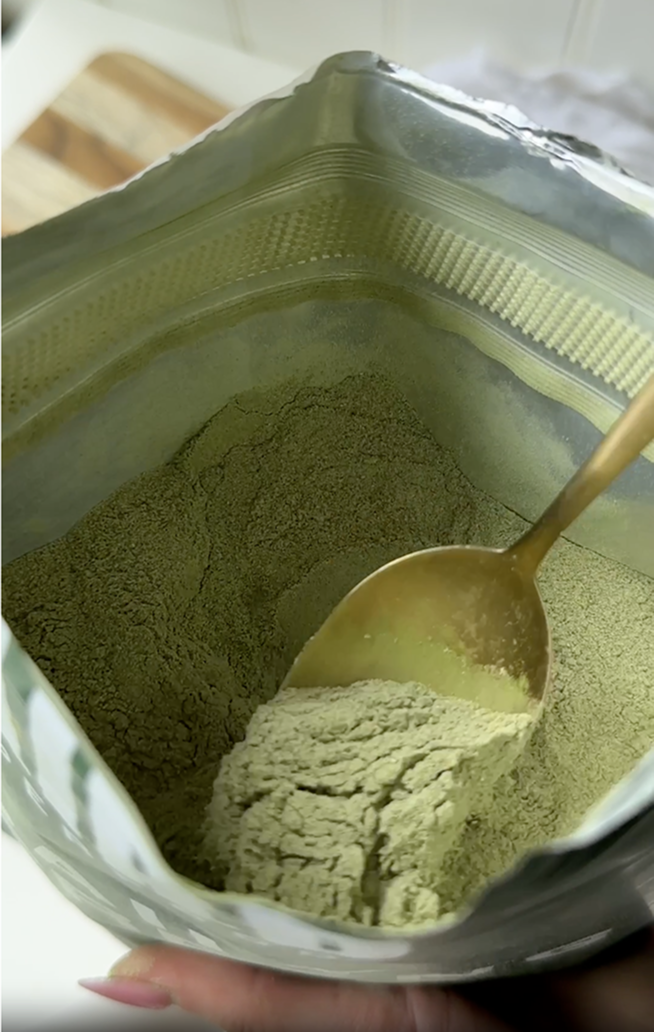 A close-up of a gold spoon scooping a fine green powder from inside a foil bag.