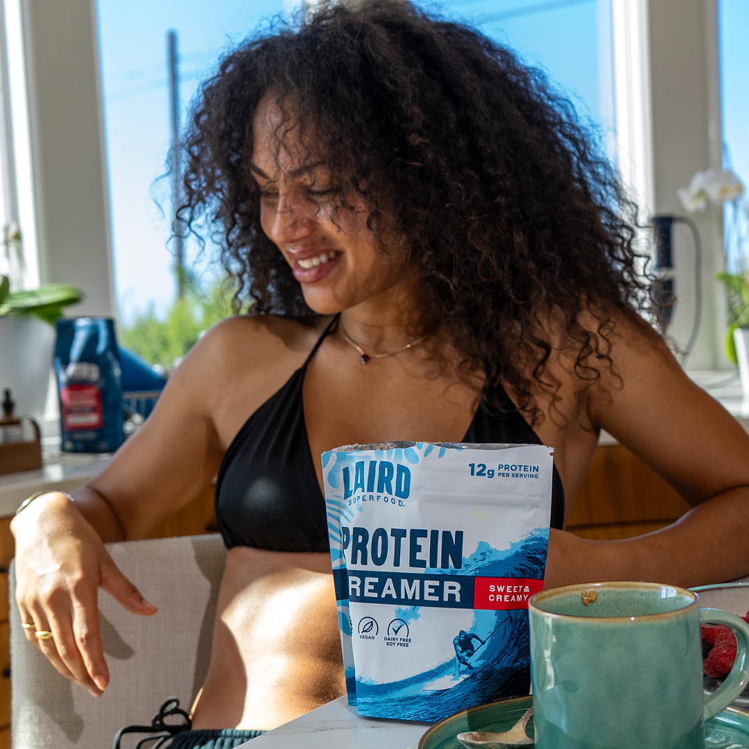 A smiling woman in a bikini top sits at a table with a bag of Laird Superfood Protein Creamer.