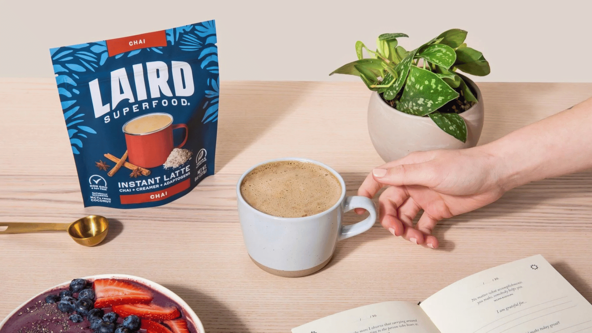 A hand reaches for a mug of chai latte next to a bag of Laird Superfood mix.