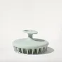 A light green, circular scalp massager with a handle, set against a plain white background.