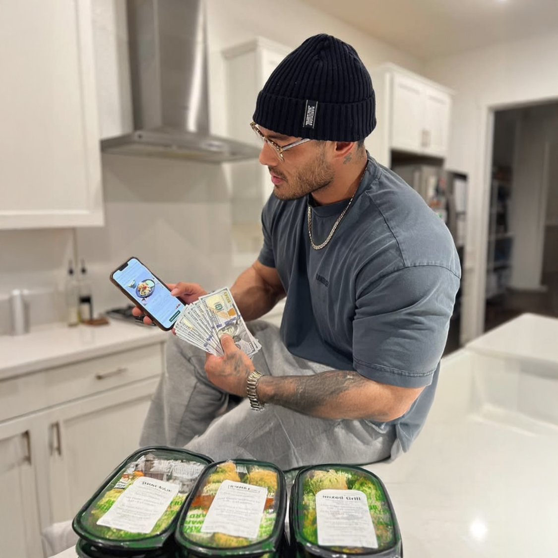 Man holding cash and phone, sitting on kitchen counter with meal packages.