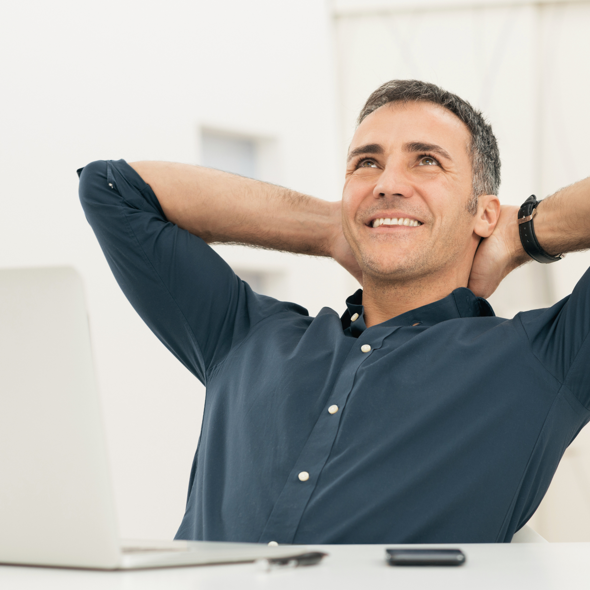A smiling man in a blue shirt leans back from his desk with his hands behind his head.