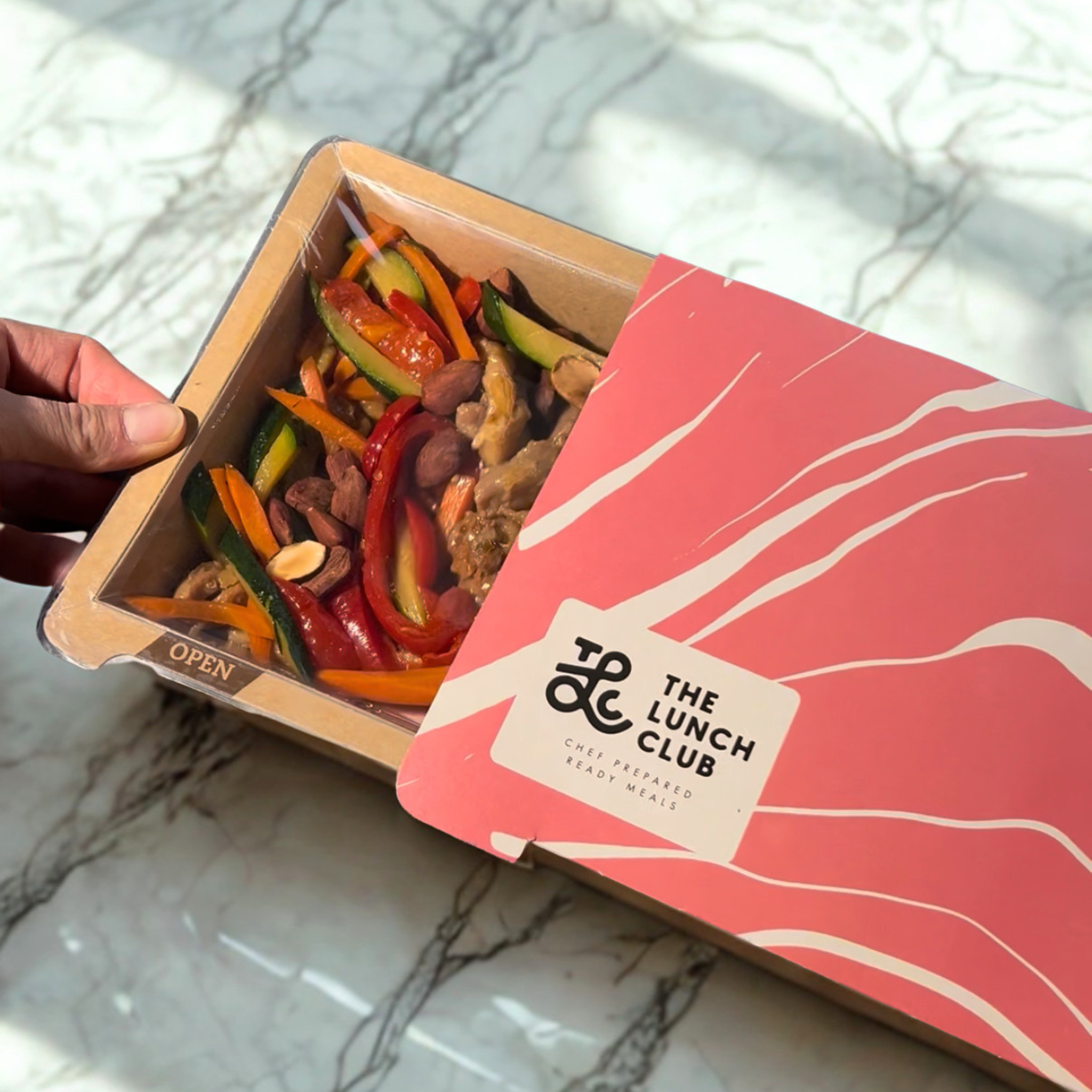 A hand opens a 'The Lunch Club' ready meal container filled with vegetables and nuts.