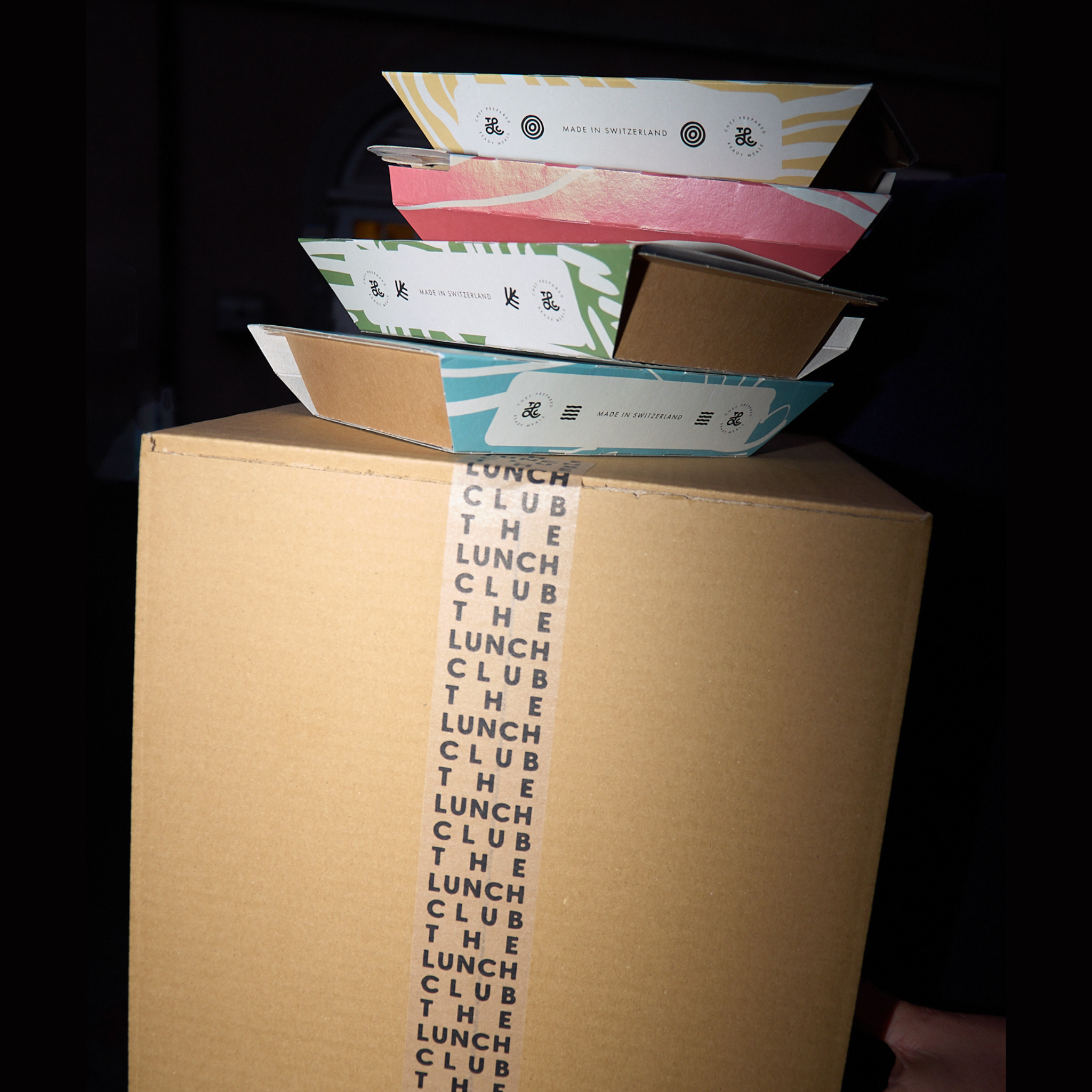 A stack of colorful meal prep boxes sits atop a larger cardboard box sealed with 'THE LUNCH CLUB' tape.