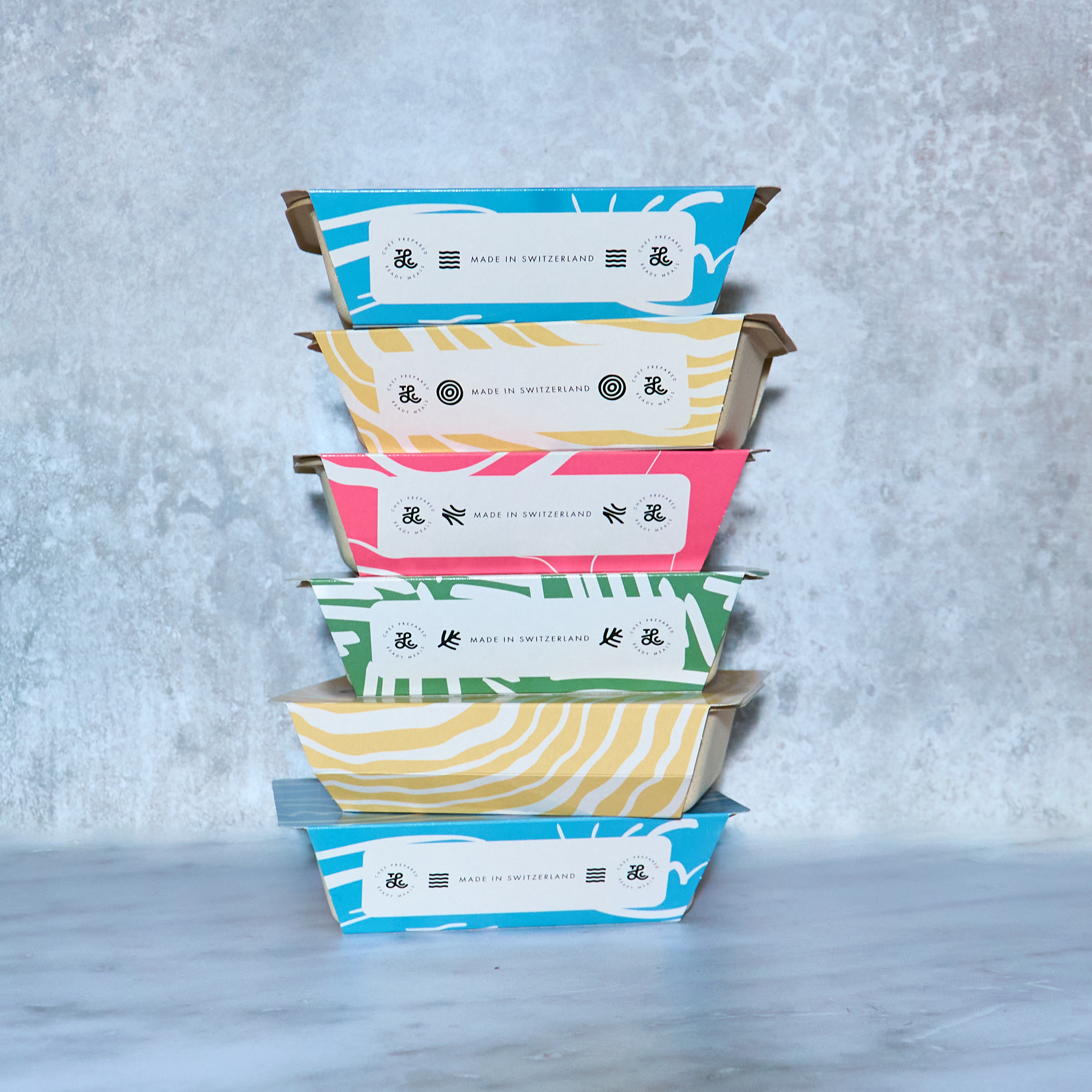 A stack of six colorful food containers, labeled 'Made in Switzerland,' against a gray marbled background.