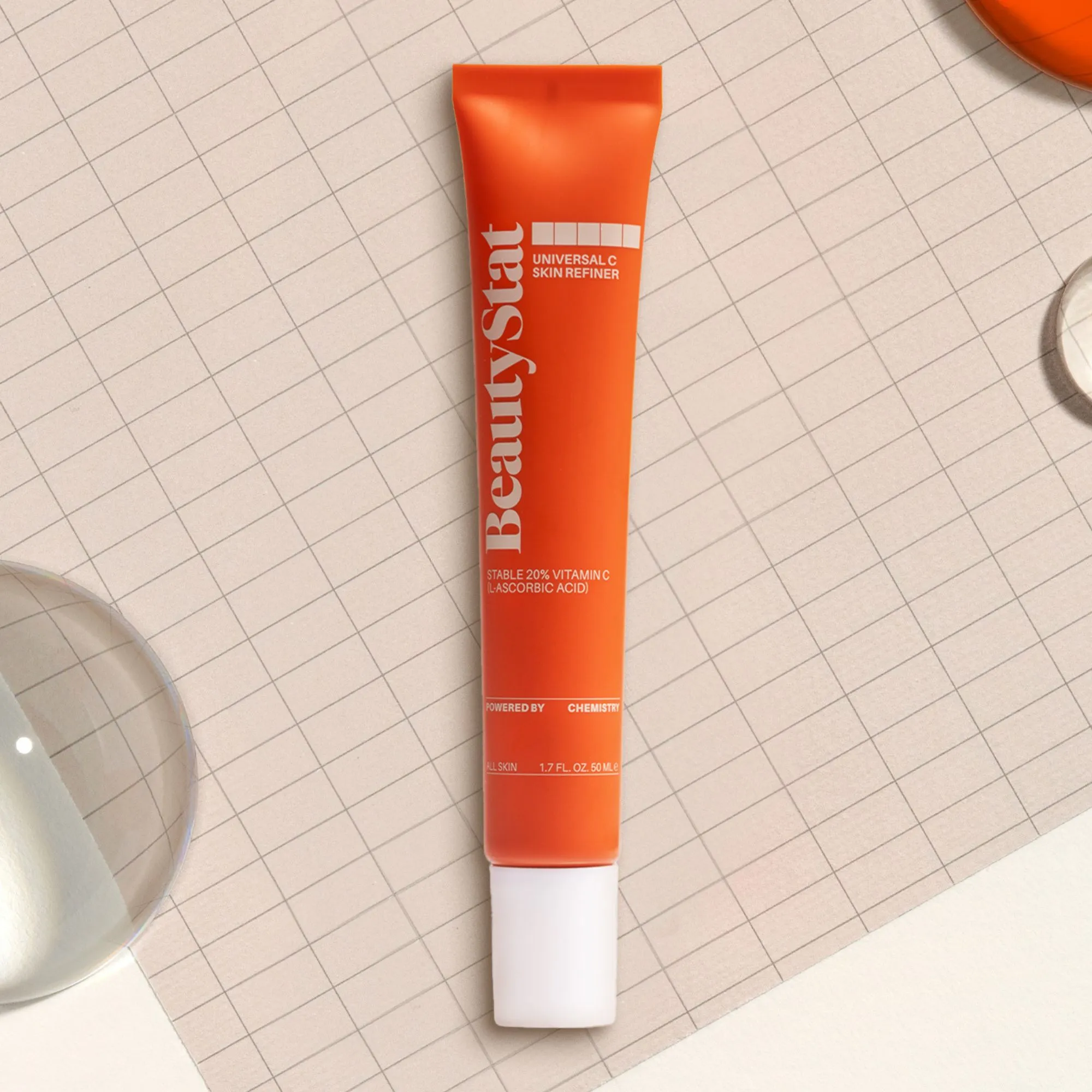 An orange tube of BeautyStat Universal C Skin Refiner lying diagonally on a gridded surface.