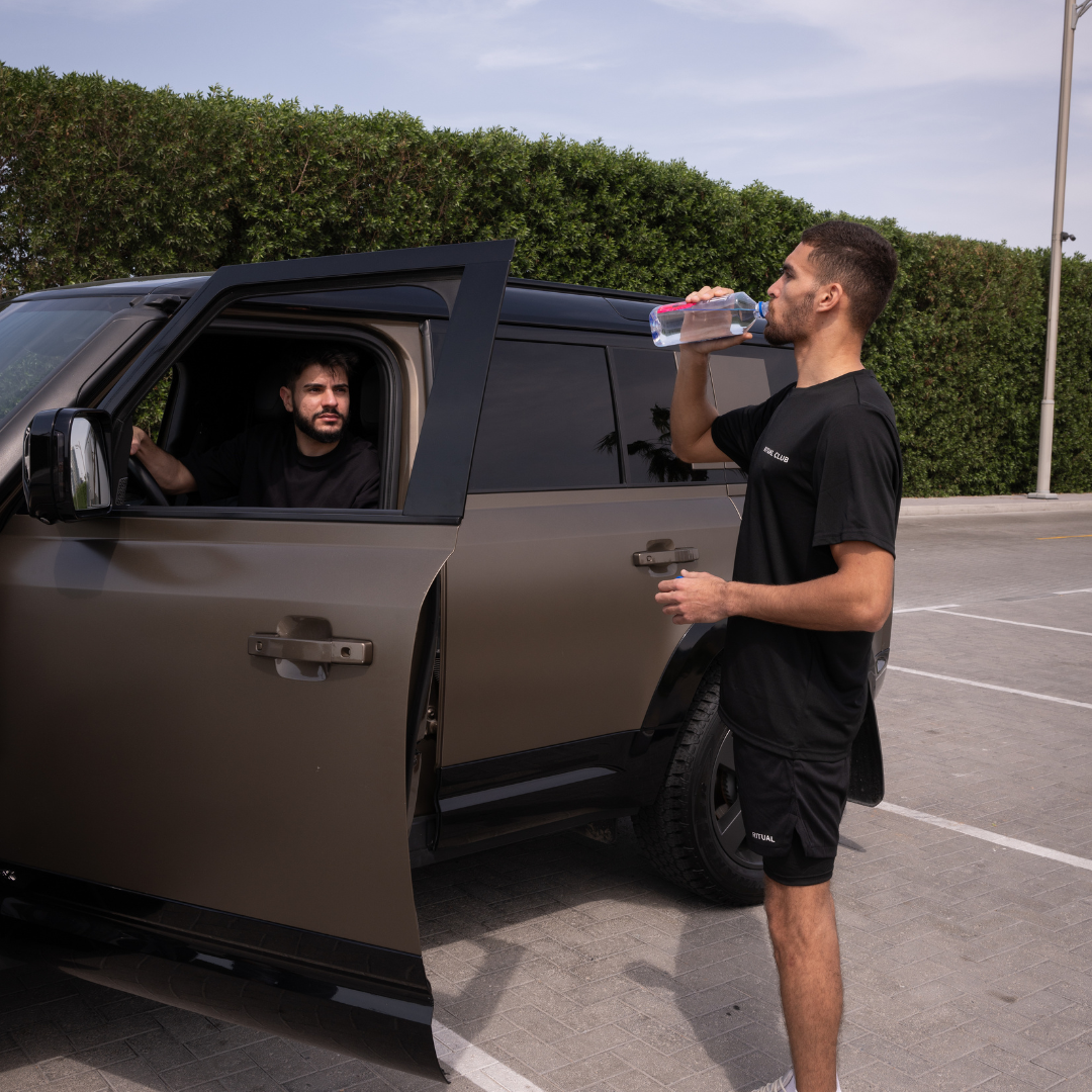 Man drinking water beside a parked SUV with another man inside.