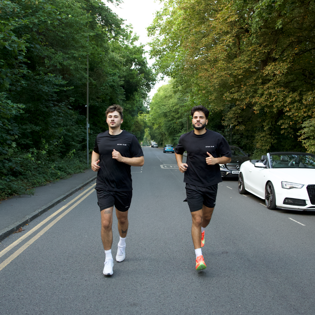 Two men jogging on a tree-lined road with parked cars.