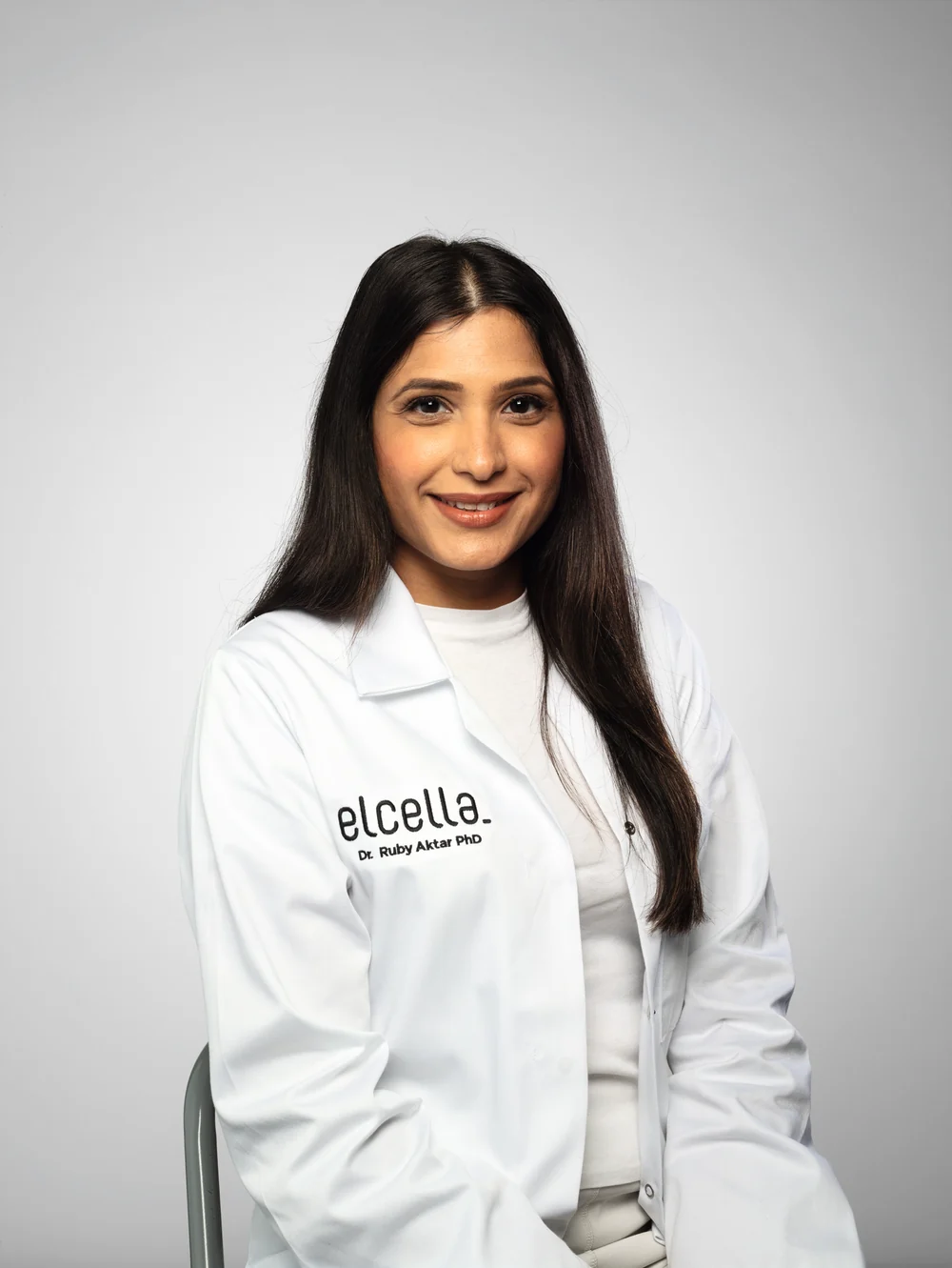 Dr. Ruby Aktar PhD smiling in a white lab coat against a light grey background.