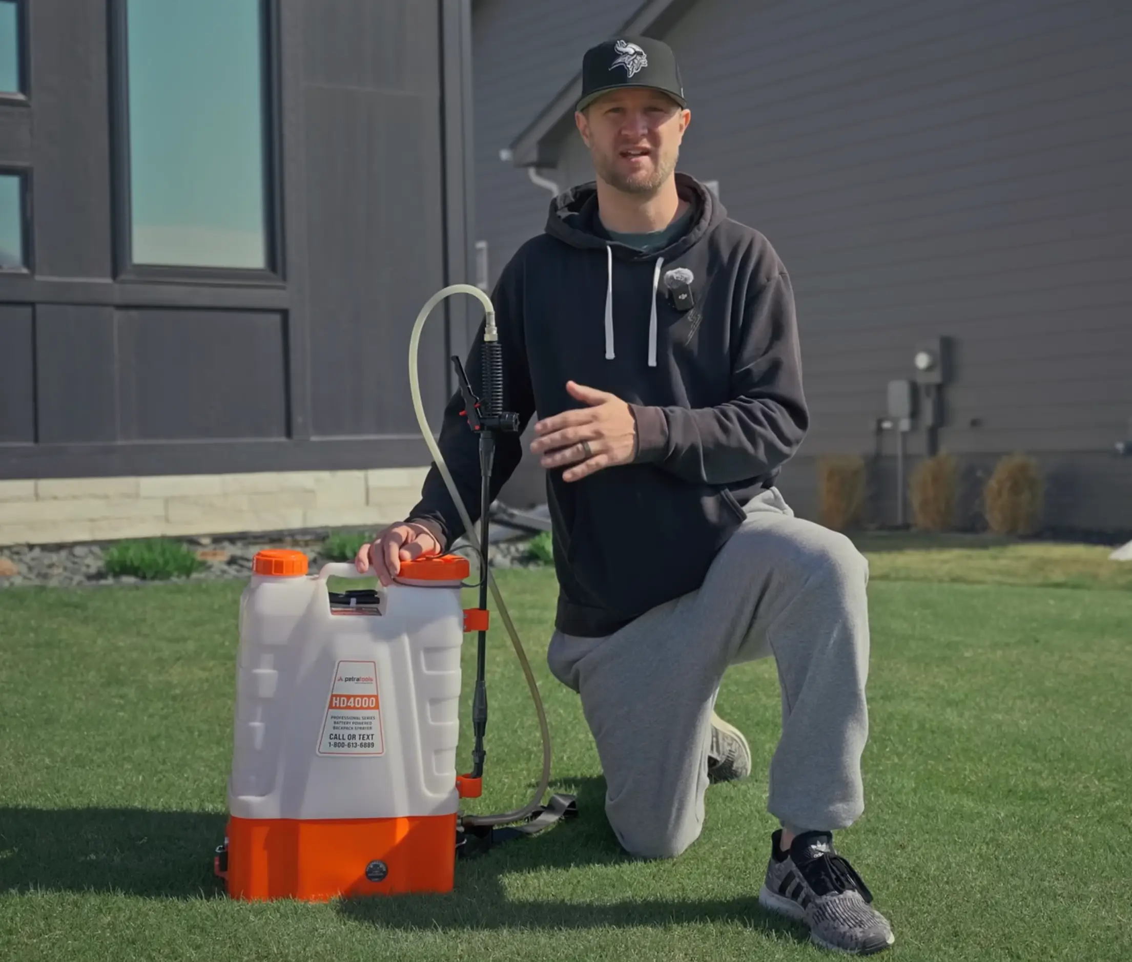 A man kneels on a lawn next to a white and orange backpack sprayer in front of a house.