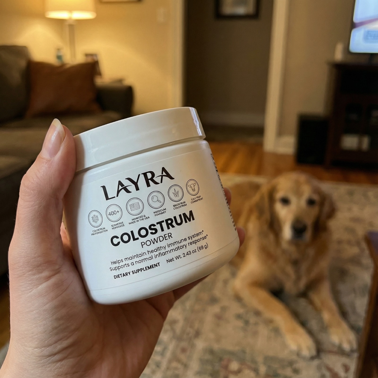 A person holds a jar of Layra Colostrum Powder, with a golden retriever blurred in the background.