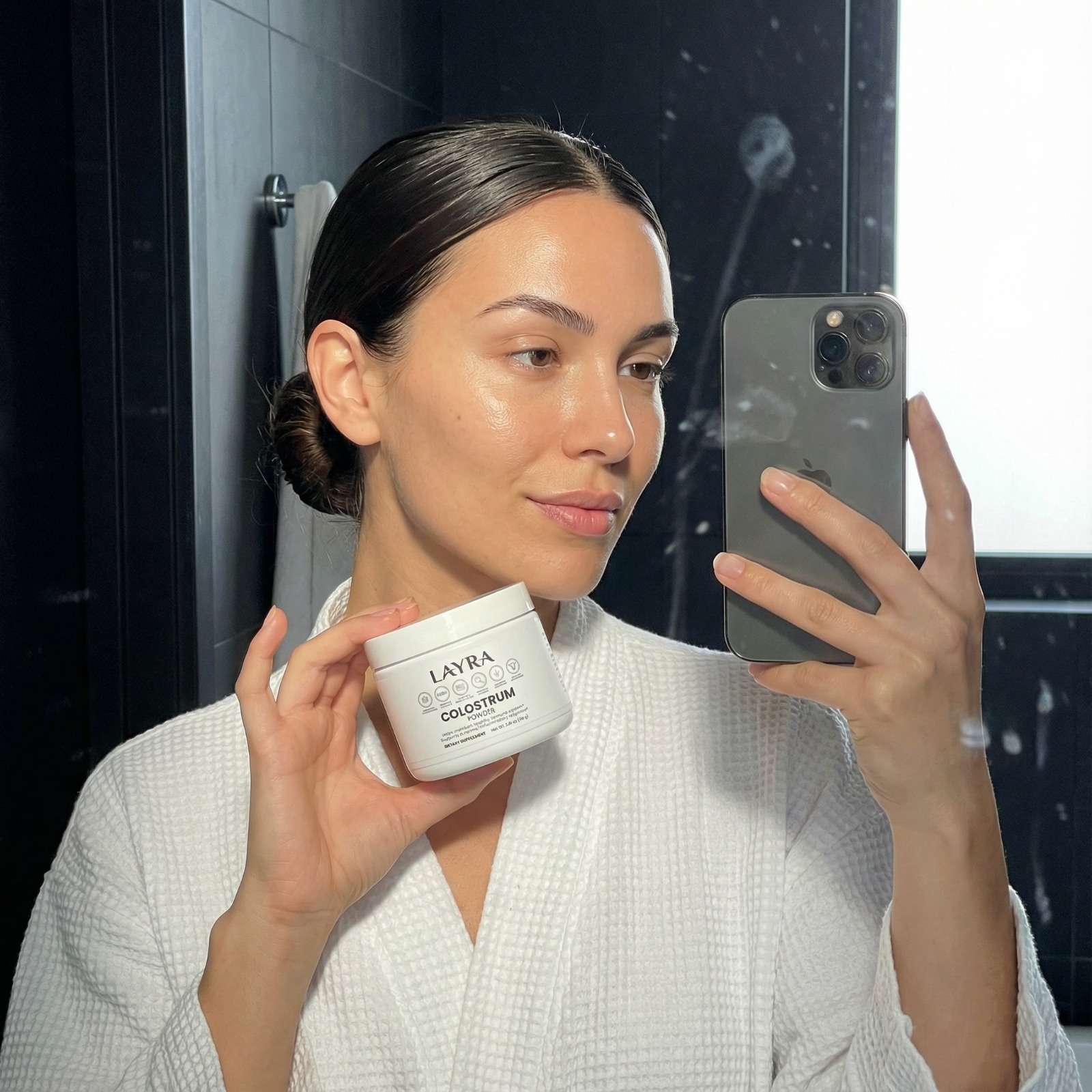 A woman in a white robe takes a mirror selfie holding a jar of Layra Colostrum Powder.
