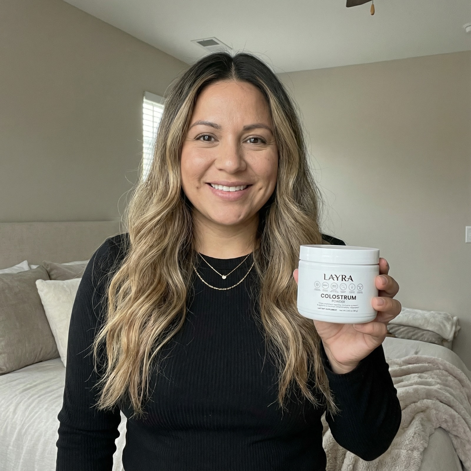 A woman smiles while holding a white jar of Layra Colostrum Powder in a bedroom setting.