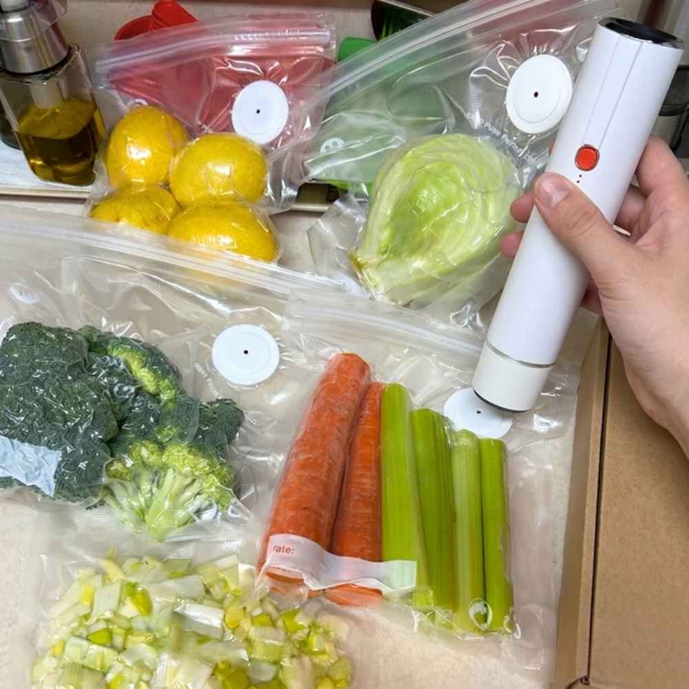 A person uses a handheld vacuum sealer on plastic bags filled with fresh vegetables and lemons.