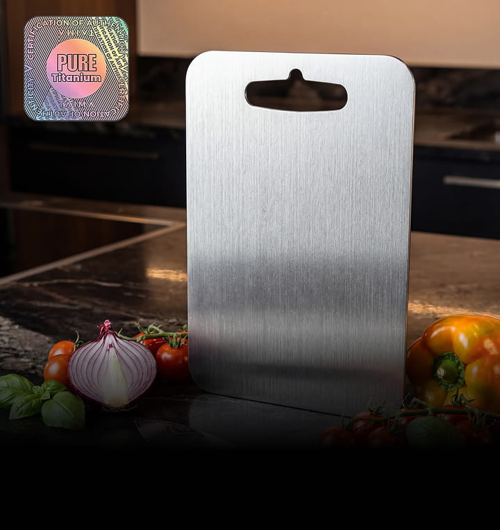 A brushed titanium cutting board stands on a dark countertop surrounded by fresh vegetables like onions and tomatoes.