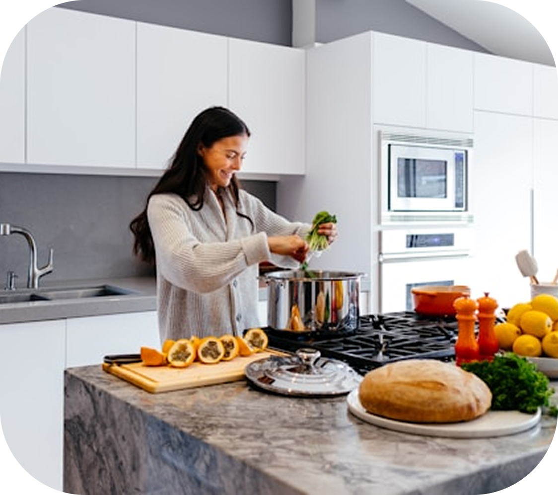 A smiling woman adds greens to a pot on the stove in a modern kitchen.