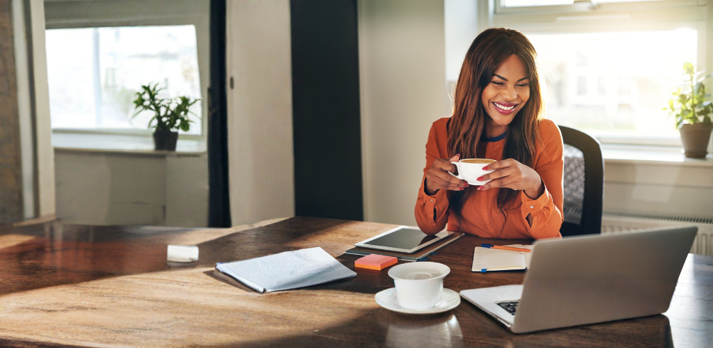 A smiling woman holds a coffee cup while sitting at a wooden table with a laptop.
