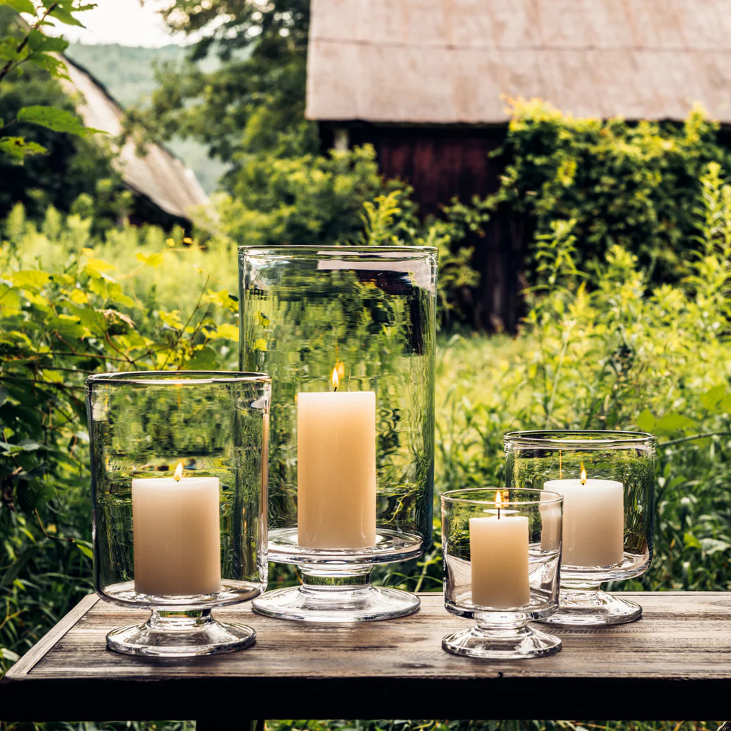Four lit pillar candles in glass hurricane holders on a wooden table in a rustic garden setting.