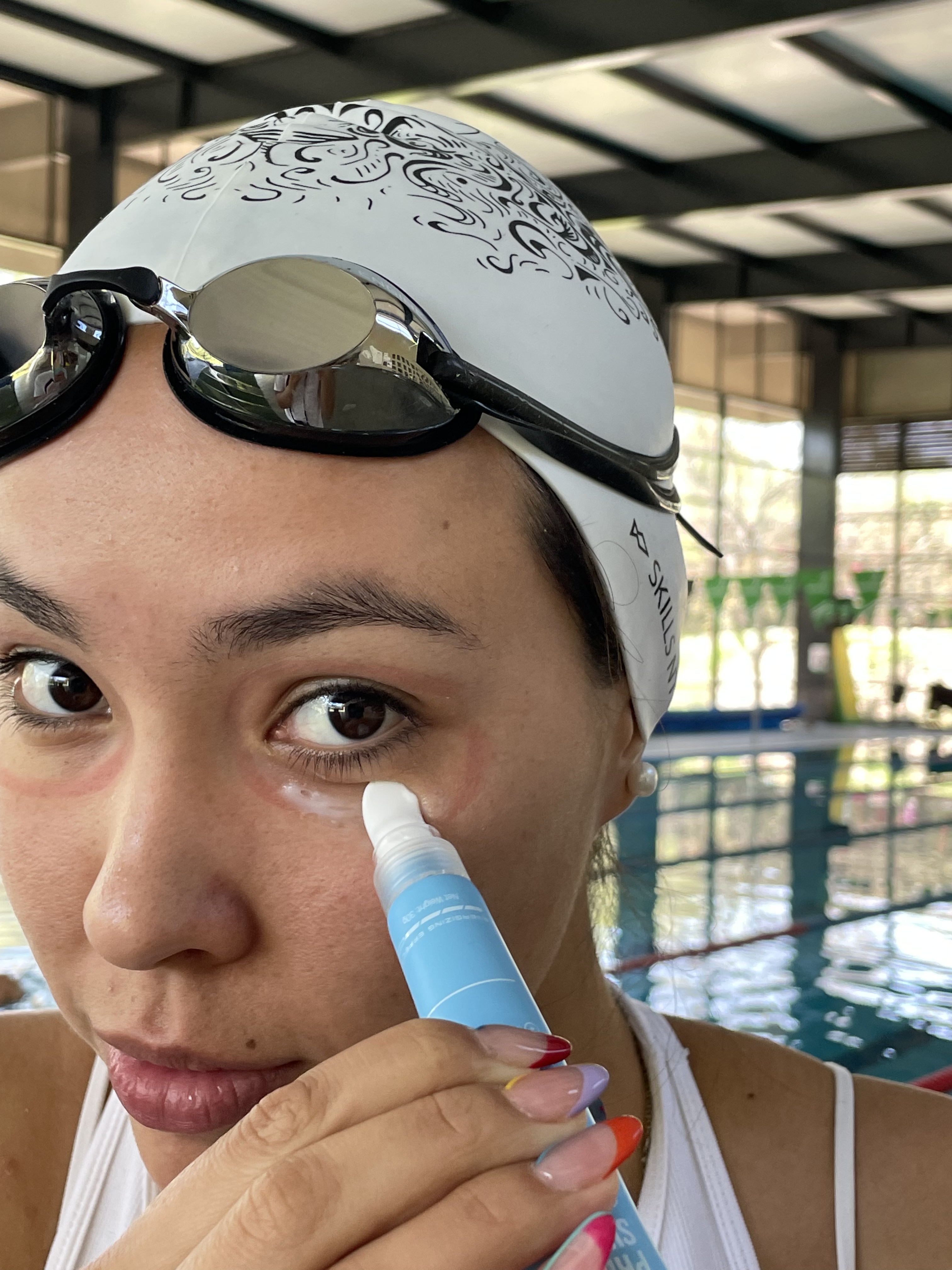 A woman in a swim cap and goggles applies a product under her eye beside a swimming pool.