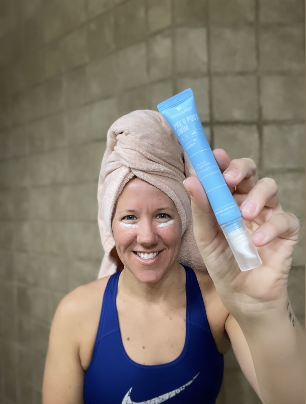 A smiling woman with a towel on her head holds up a blue tube of eye gel.
