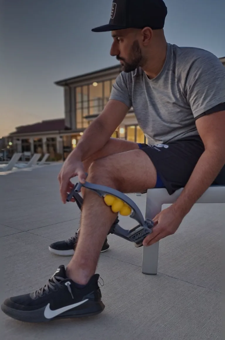 A man in athletic gear sits on a bench outdoors using a roller massager on his lower leg.