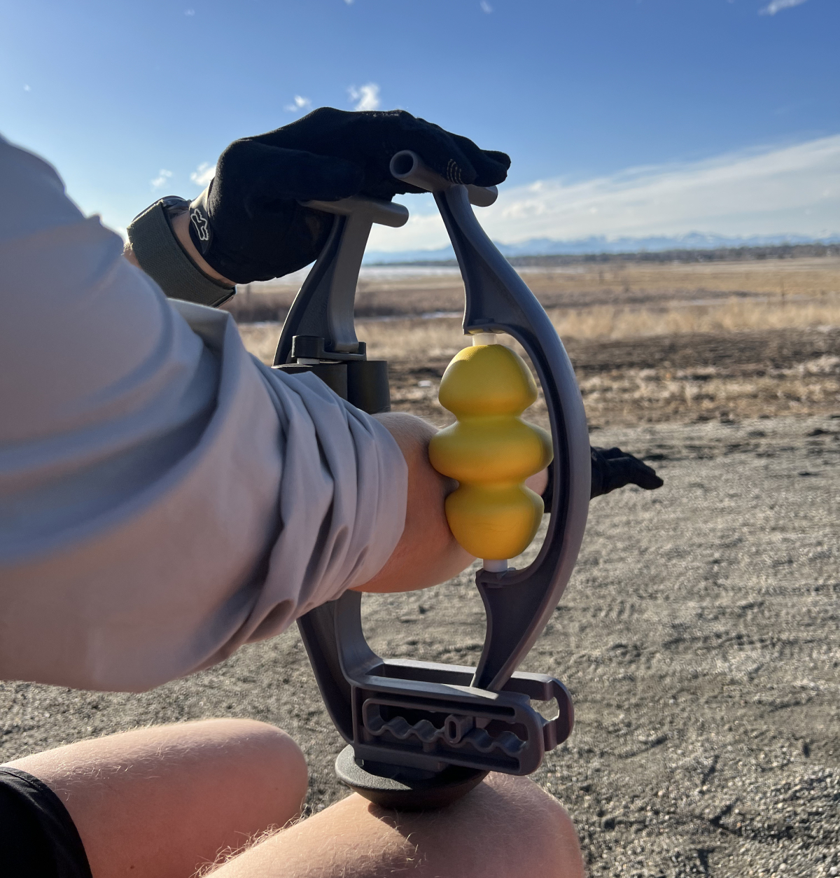 A person in gloves uses a handheld massage tool with a yellow roller on their forearm outdoors.