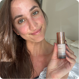 A woman with long brown hair smiles while holding up a small bottle of a beauty product.