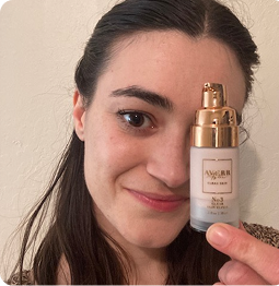 A smiling woman holds a small bottle of a beauty product next to her eye.