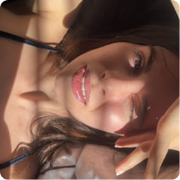 A close-up of a woman lying down, smiling at the camera with sunlight and shadows on her face.