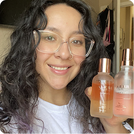 A smiling woman with dark curly hair and glasses holding two small bottles of liquid product.