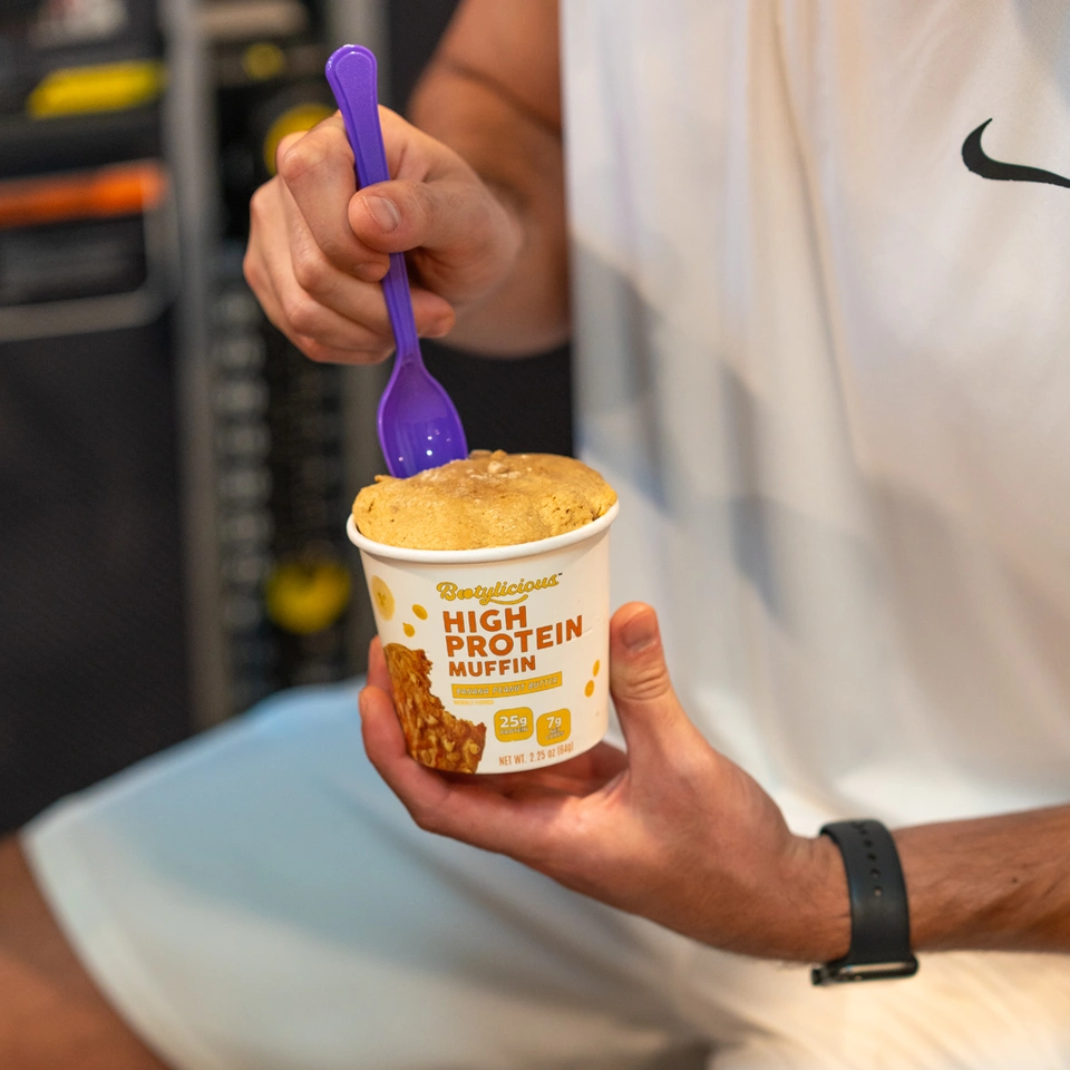 A person holds a cup of Bootylicious High Protein Muffin with a purple spoon inserted in it.