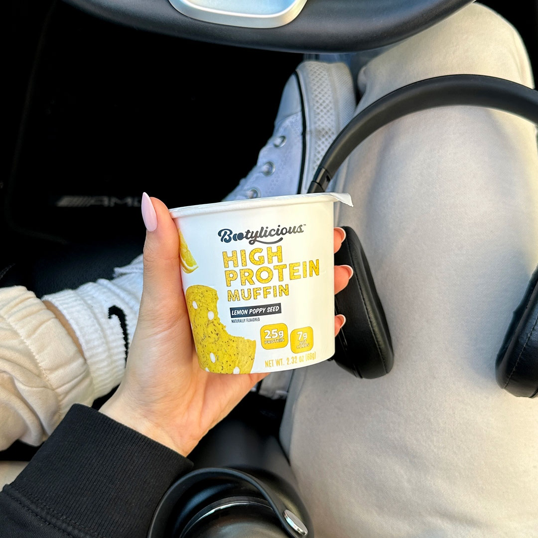 A person in a car holds a cup of Bootylicious High Protein Lemon Poppy Seed Muffin.