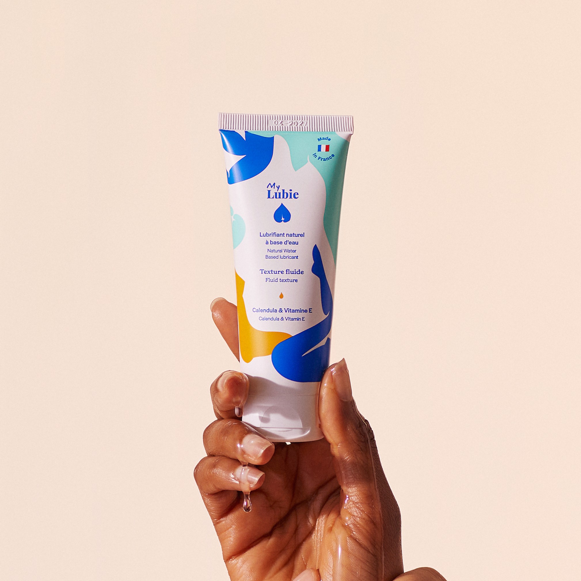 Hand holding a colorful tube of lubricant against a beige background.