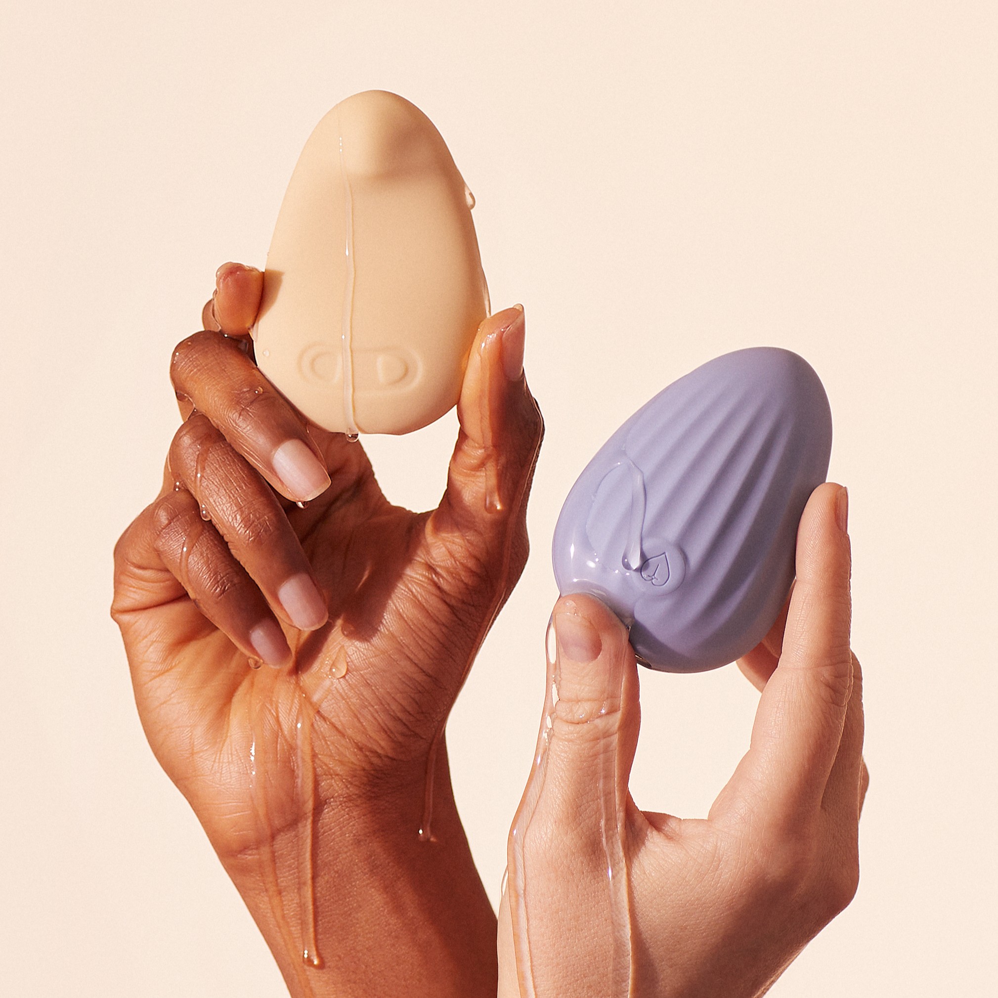 Two hands holding beige and purple egg-shaped objects with water droplets.