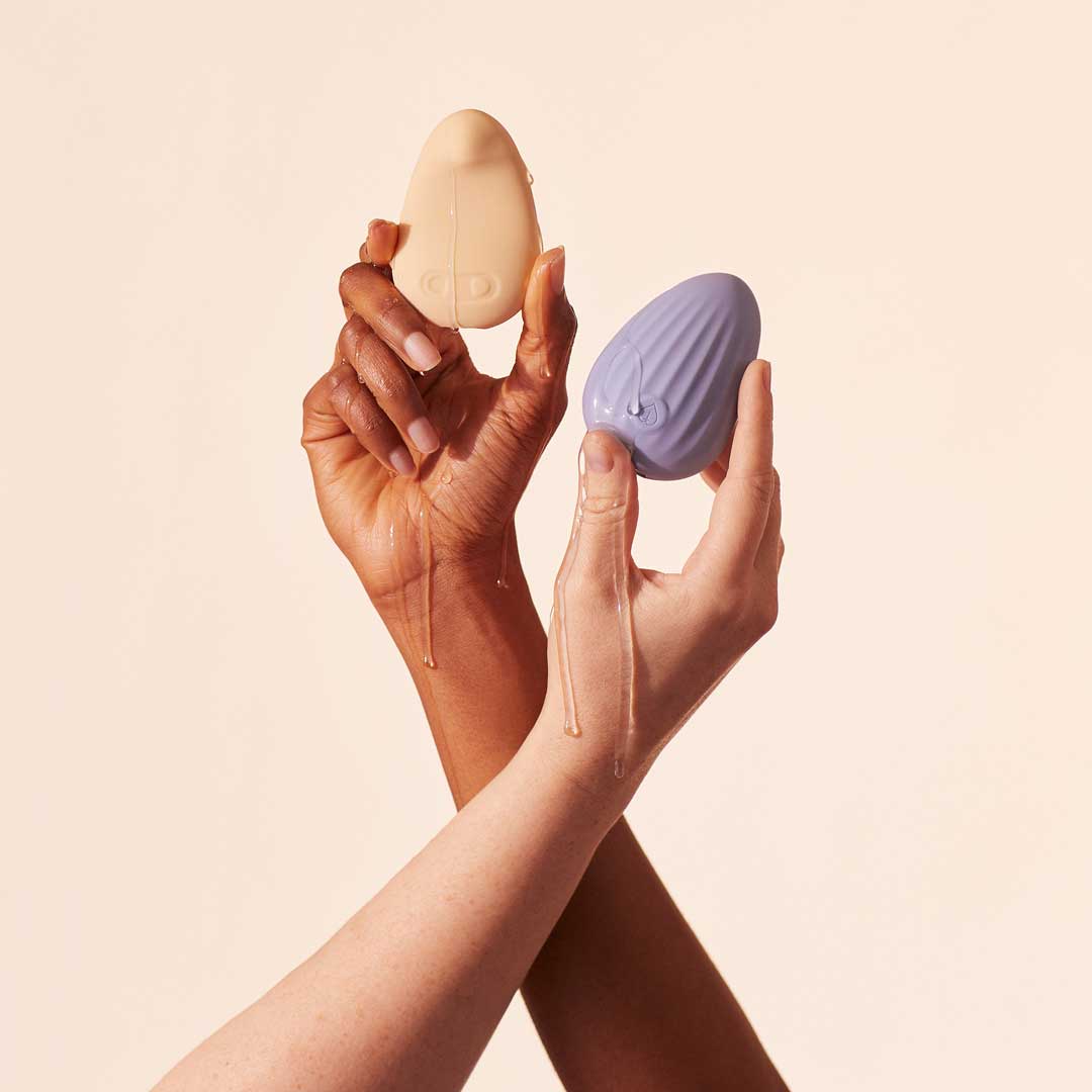 Two hands holding beige and purple egg-shaped objects with liquid dripping.